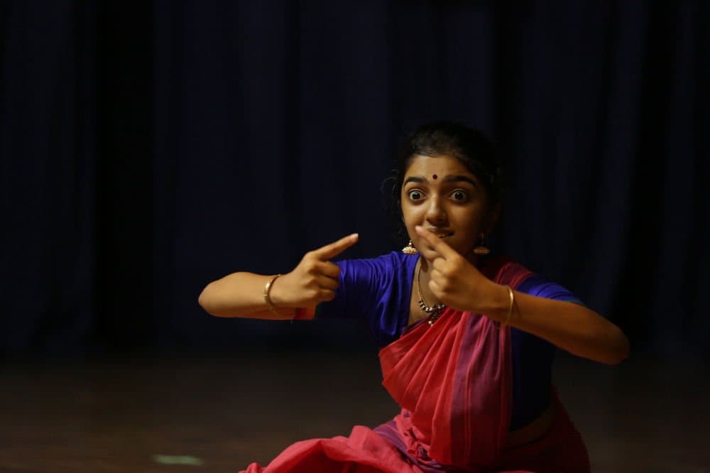 A young dancer in a blue blouse and red skirt on a dark stage, hands raised beside her face in an expressive pose